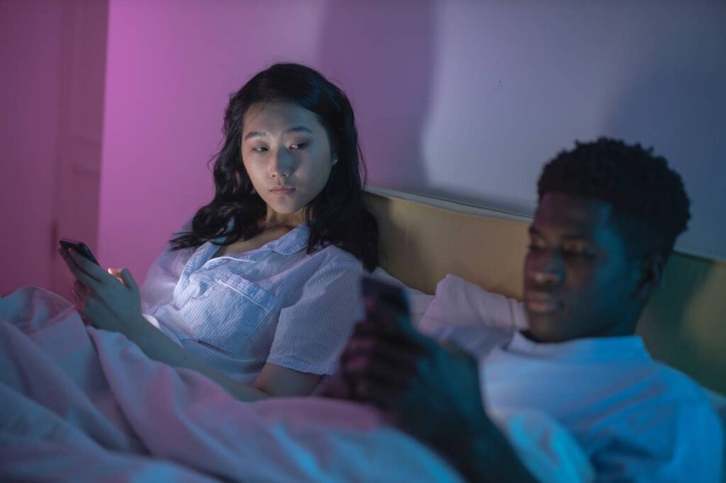 En una cama, una pareja muestra sus celos mirando el móvil de la otra persona.
